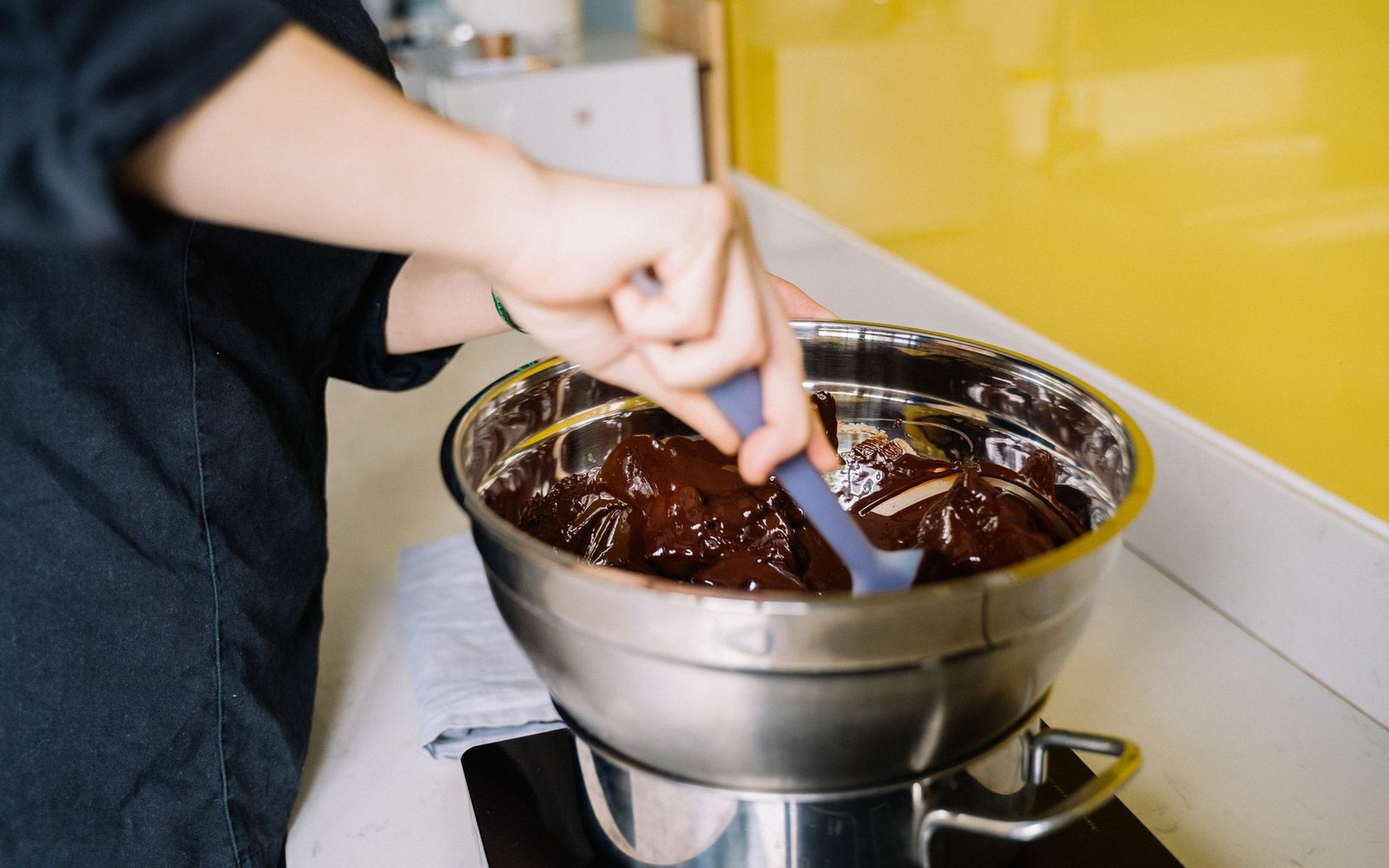 A glass bowl of dark ganache being emulsified with a rubber spatula, the ribbon trail visible on the surface, shot from above on a dark wooden bench