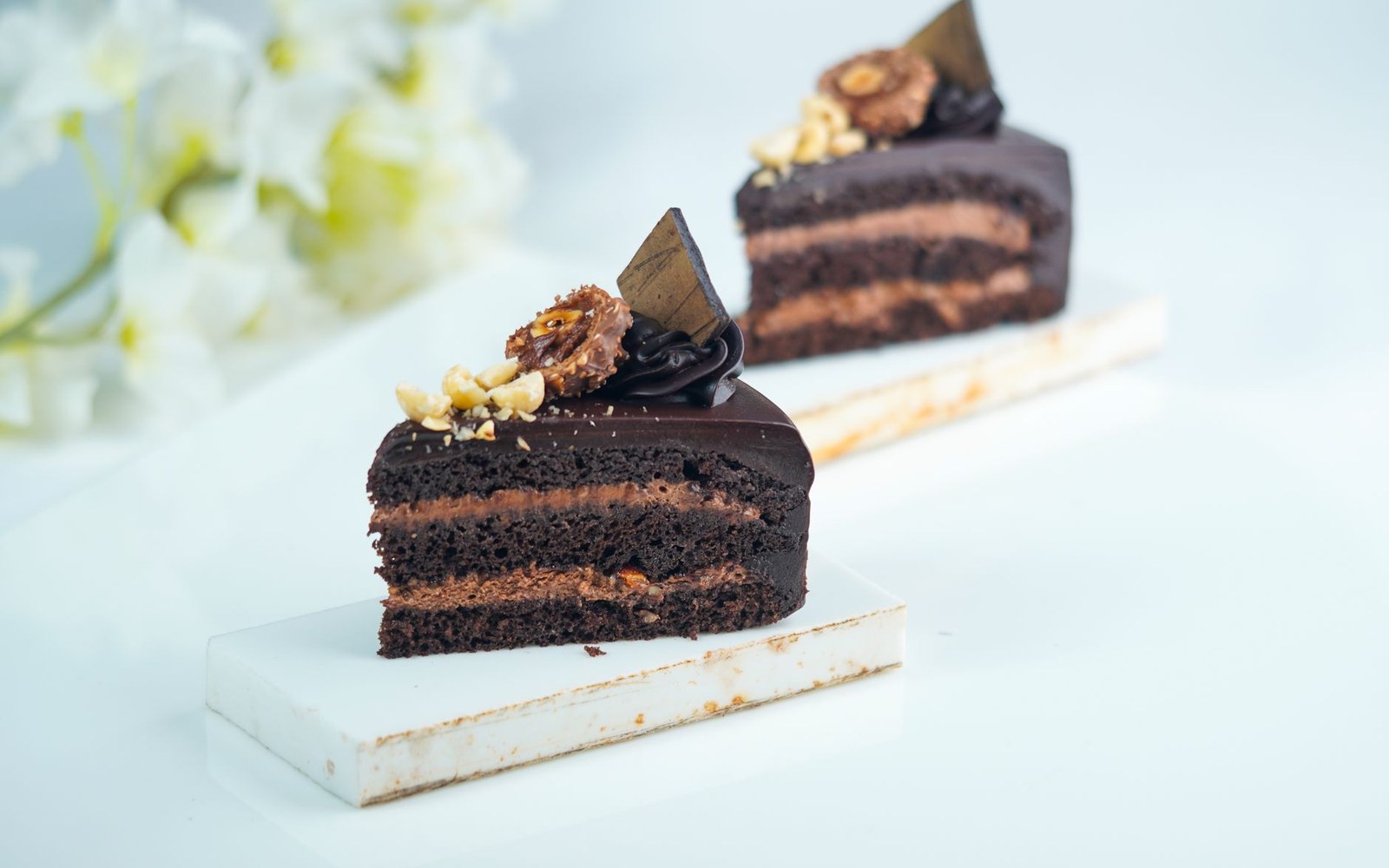 A tall slice of layered chocolate cake with a ganache finish and a chocolate shard, on a white marble slab