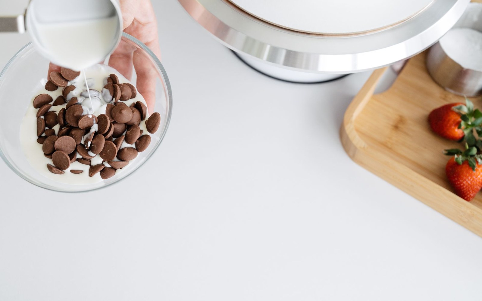 Milk being poured from a jug over chocolate callets in a glass bowl, a mixer bowl and strawberries behind