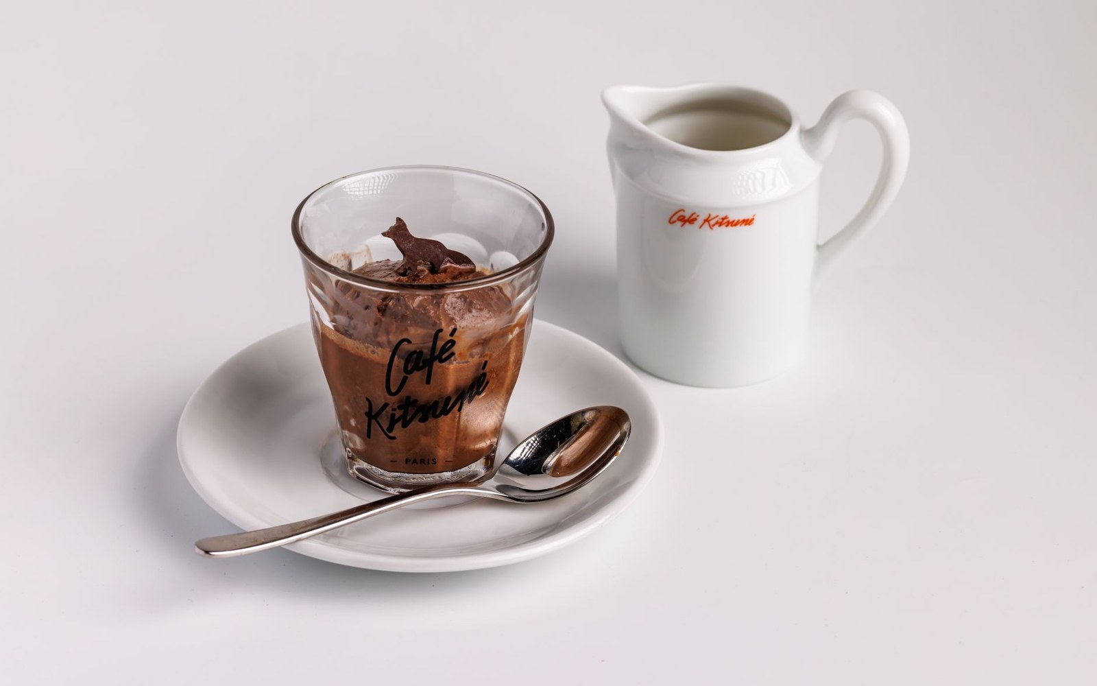 A glass of chocolate mousse on a saucer with a spoon and a small white jug of cream