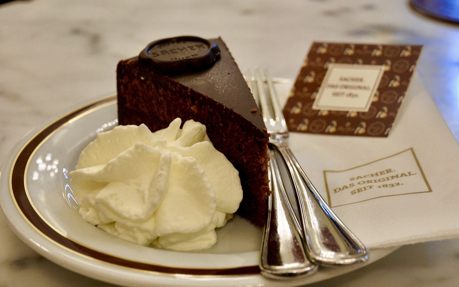 A slice of Sachertorte carrying the Sacher seal, served with whipped cream on a marble café table