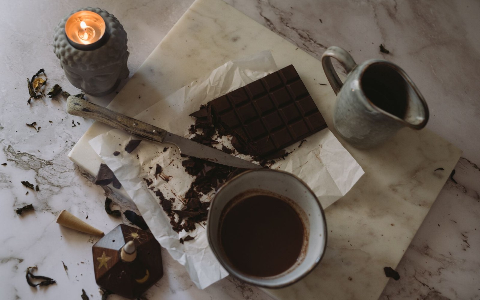A broken dark chocolate bar on marble beside a cup of drinking chocolate, a knife and a lit candle, shot from above