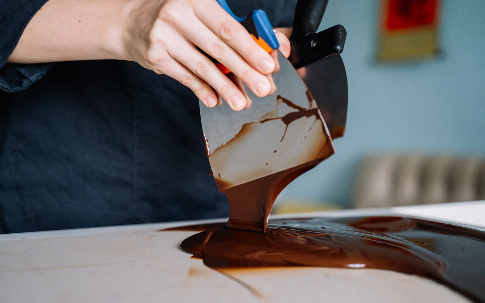 An adult pastry cook's hands working tempered chocolate across a white marble slab with a metal scraper, mid-tabling, the chocolate glossy and viscous in the bench light